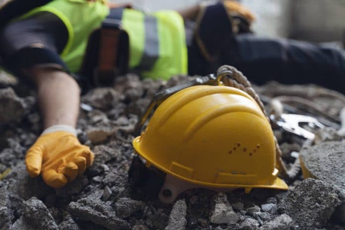 Un casque jaune repose sur les décombres à côté du bras tendu d'un ouvrier du bâtiment portant un gilet de sécurité et des gants, soulignant les graves conséquences d'un accident de chantier.